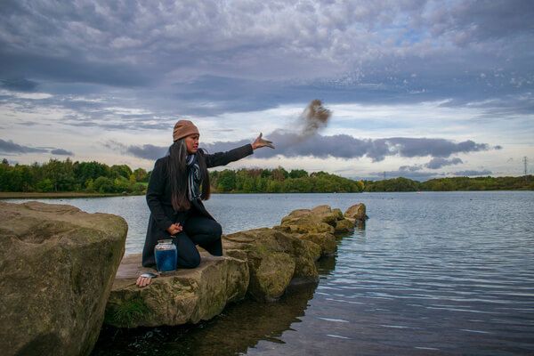 Person sitting on a rock by a lake, tossing a handful of dirt into the air under a cloudy sky.