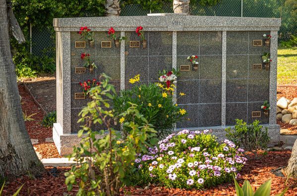 Granite memorial wall with engraved plaques, red flowers, and a bed of colorful daisies in a landscaped garden