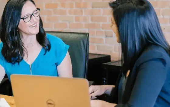 Two people talking across a table with a laptop in a brick-walled office