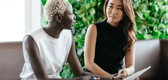 Two people in business attire talk on a couch in a bright, modern office setting.