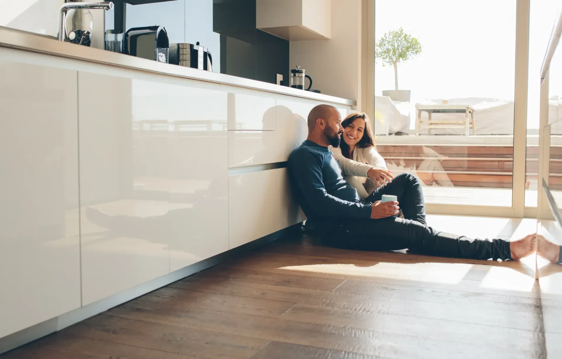 Two people sitting on a kitchen floor in warm sunlight, leaning against cabinets, with a baby nearby.
