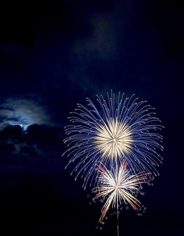 Fireworks bursting in a dark night sky, with blue and white sparks above glowing clouds.