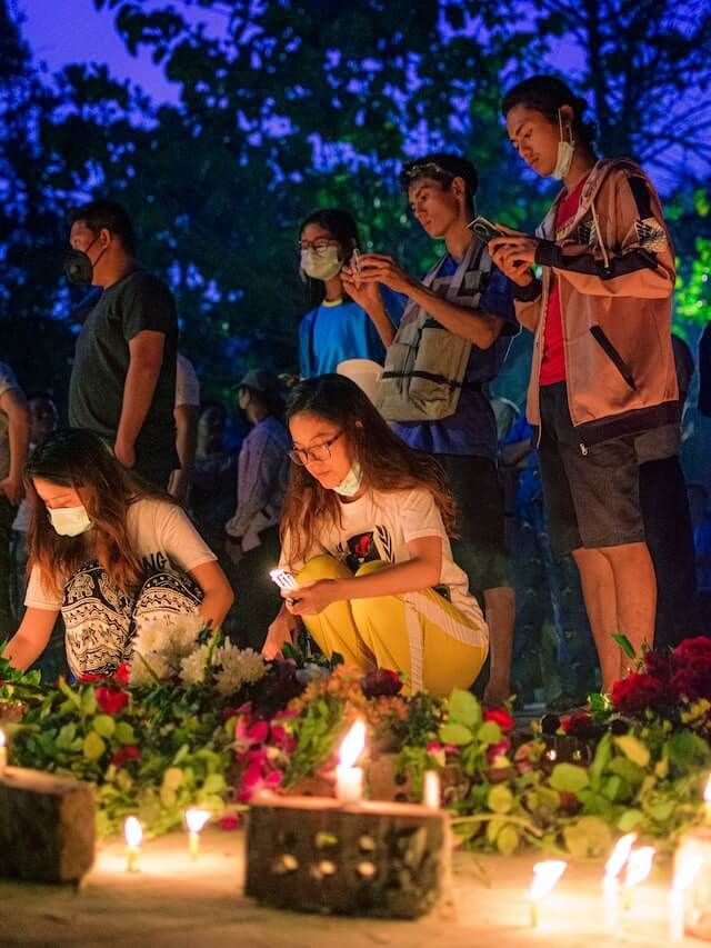 People kneeling and standing among candles and flowers at a nighttime vigil outdoors