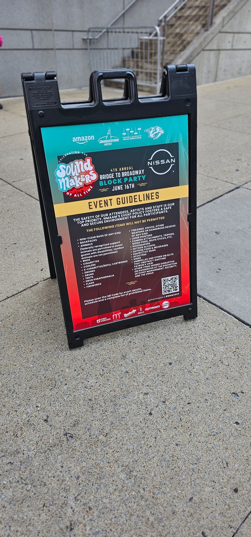 Black sandwich board sign on a sidewalk displays a menu with event information and a QR code.