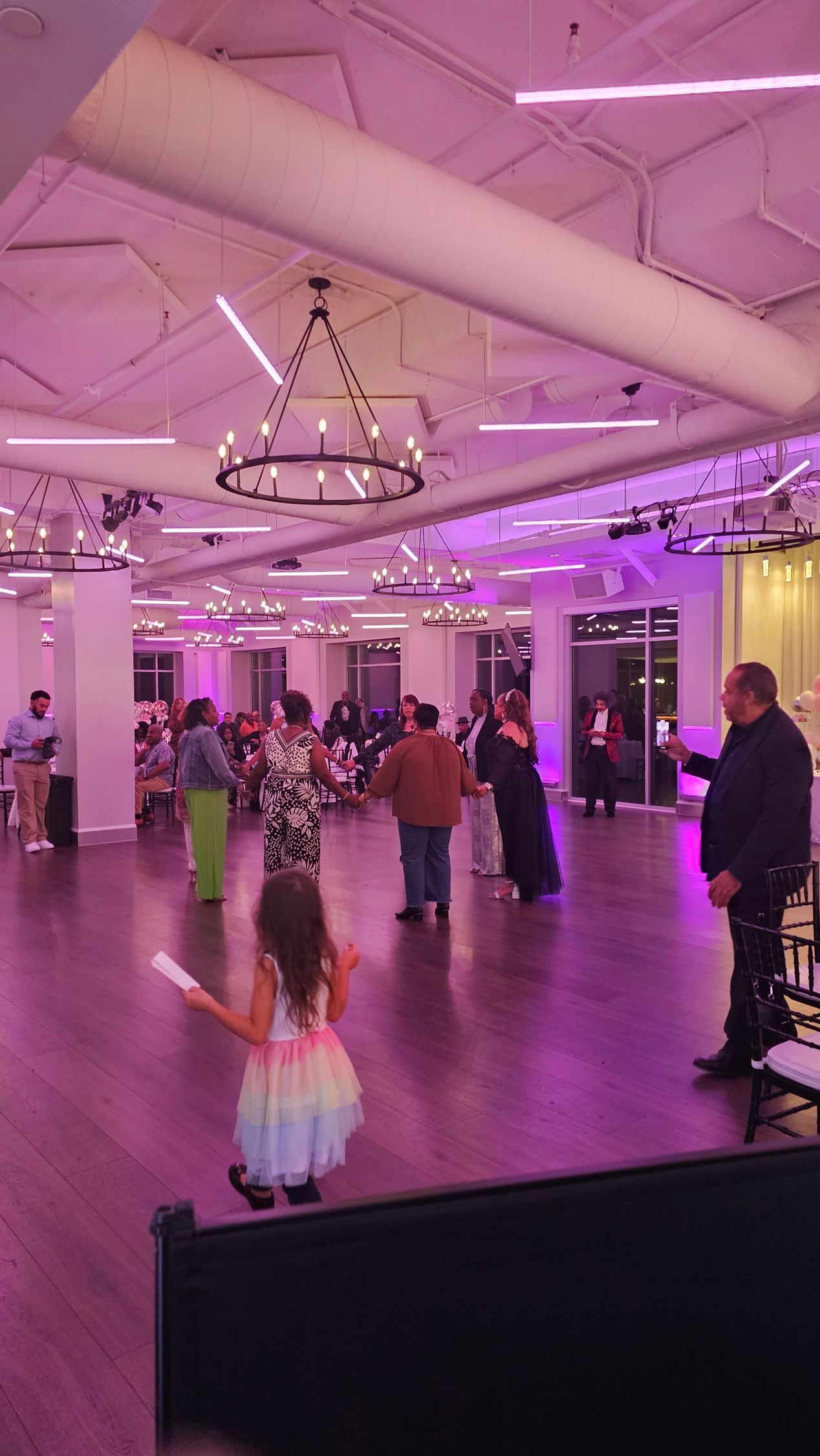 Child in colorful dress dances in ballroom with others, purple lighting.