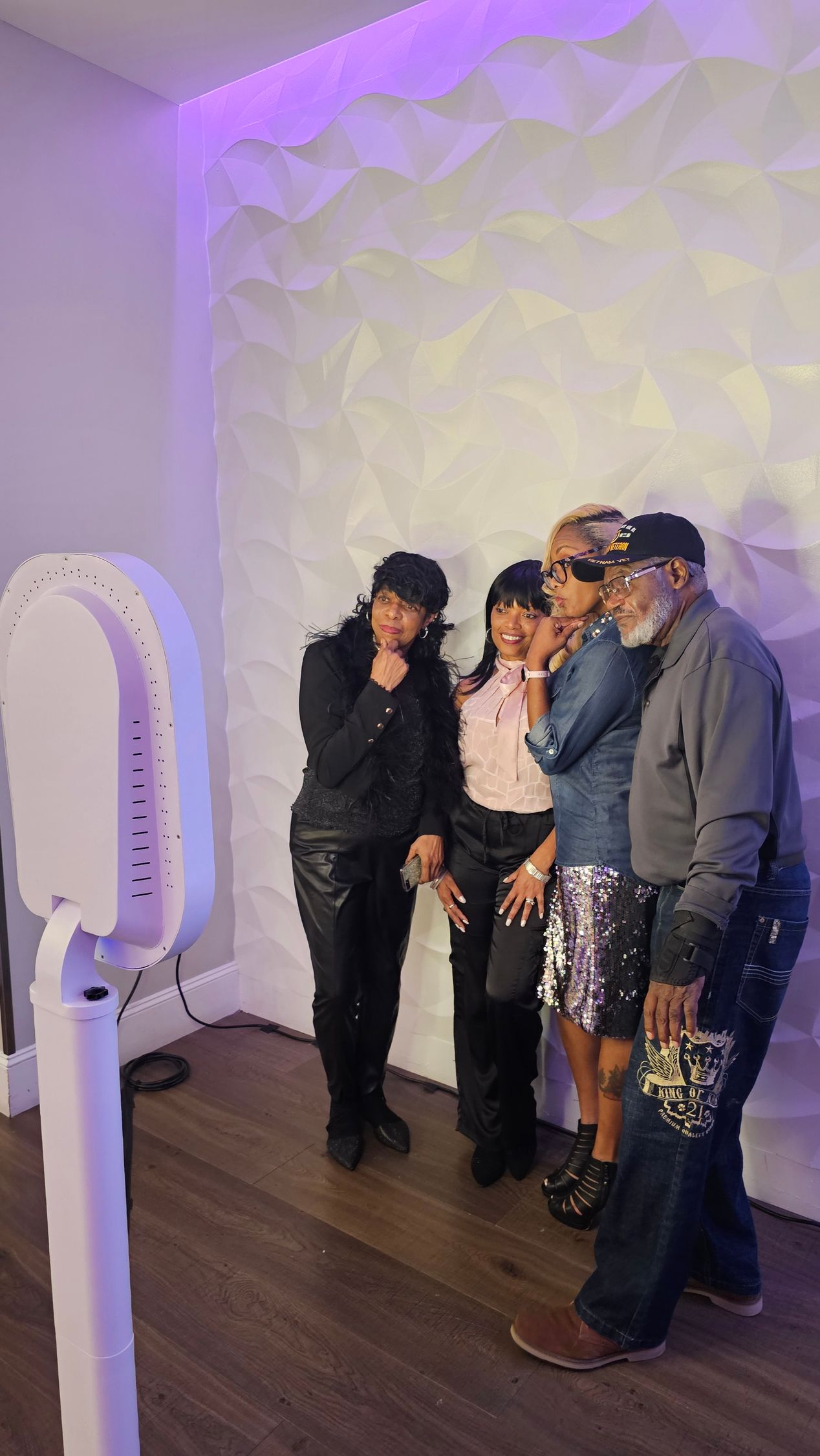 Five people pose in a photo booth with a textured white wall. They blow kisses, lit with purple light.