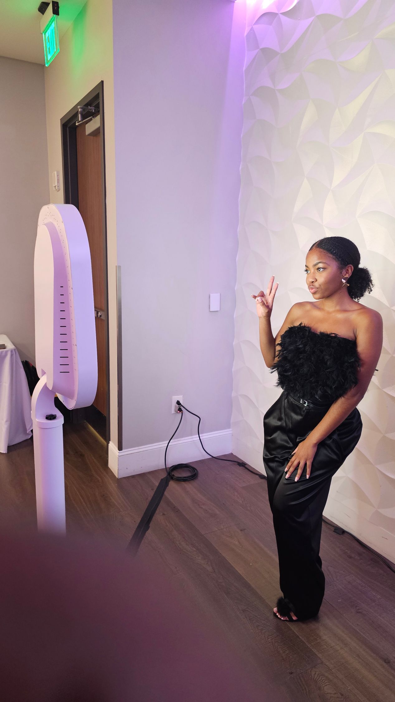 Woman poses next to photo booth. She wears a black, feathered top and pants, indoor setting with white wall.
