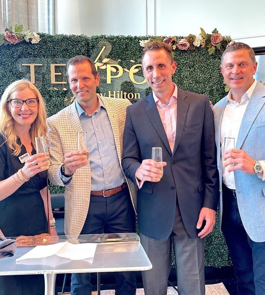 Four people, holding glasses, toast in front of a Tempo by Hilton sign. Green hedge backdrop.