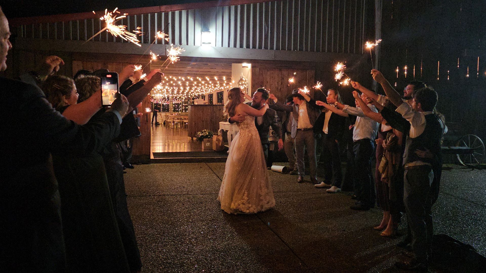 Newlyweds embrace as guests wave sparklers outside a warmly lit building at night.