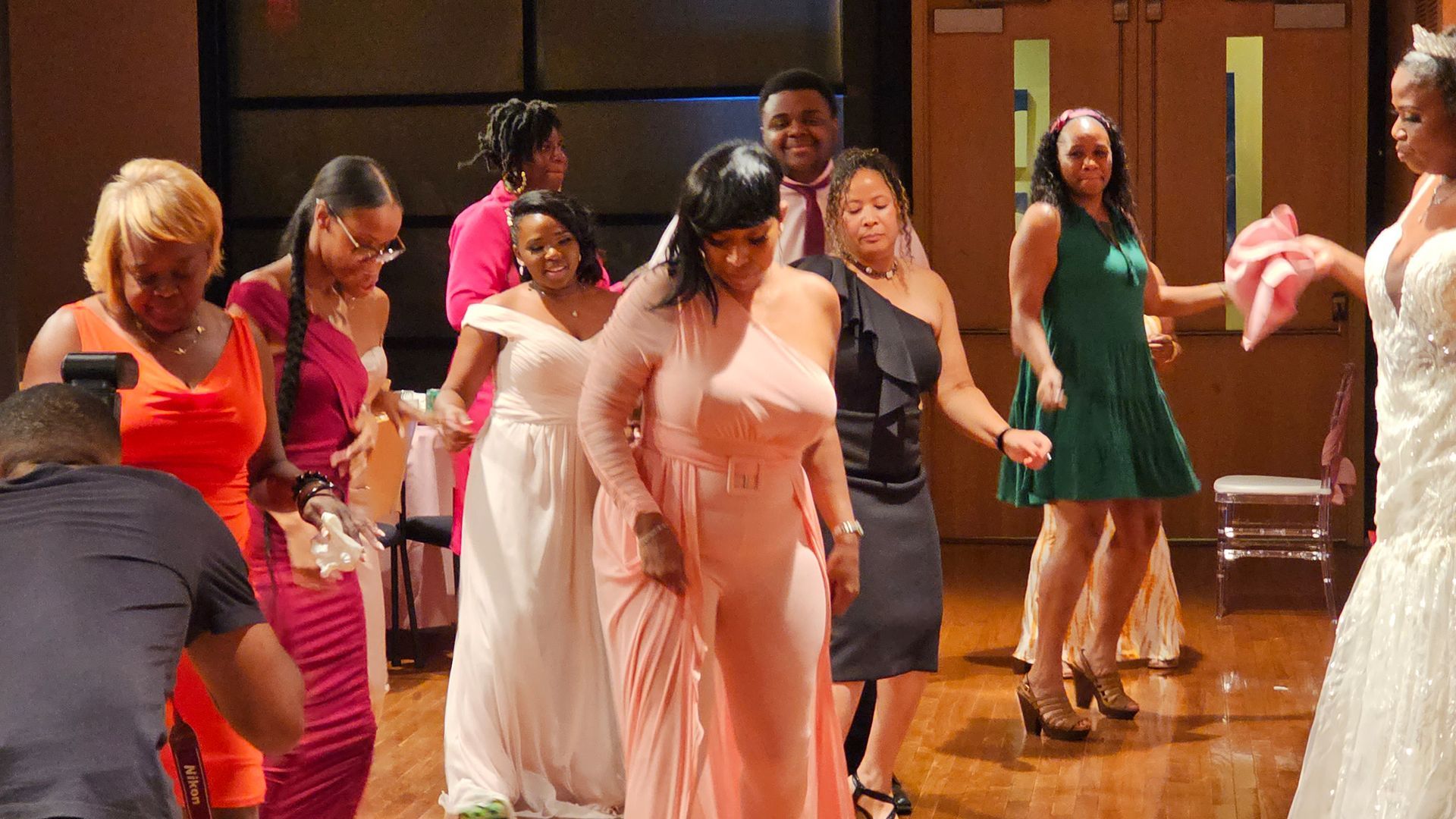 People dancing at an event, wearing gowns. Indoor setting with a wooden floor and neutral walls.
