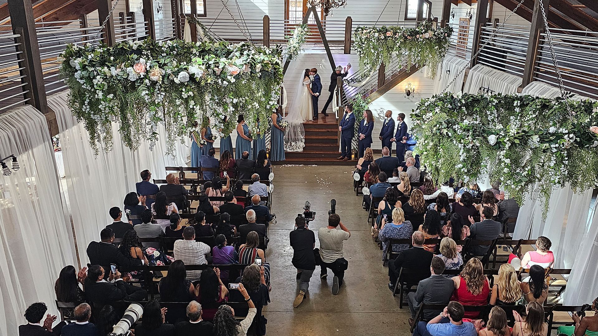 Wedding ceremony in a barn; couple on a stage under floral arches, guests seated in rows, photographer taking pictures.