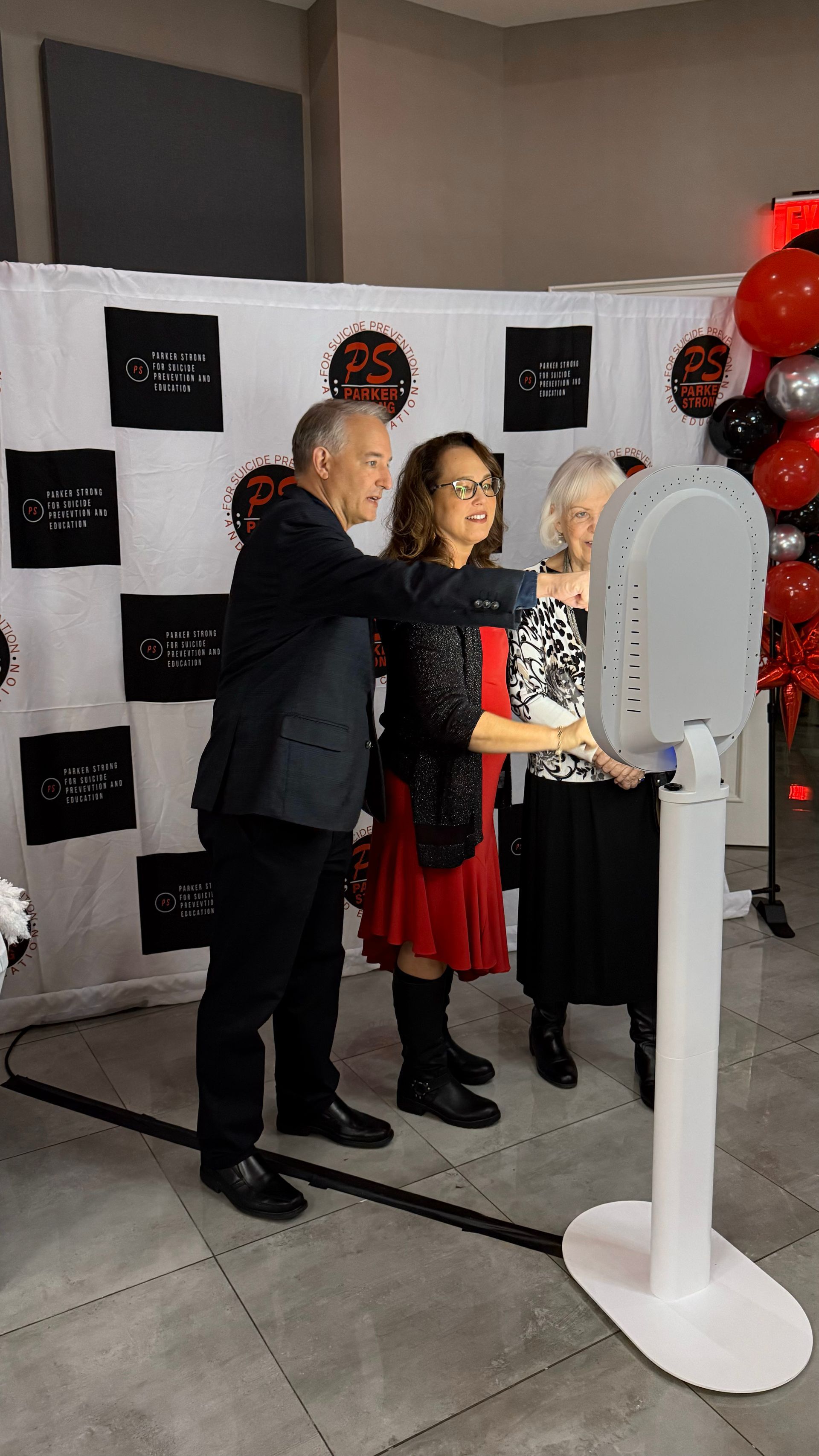Three people posing at a photo booth with a black and white backdrop and red balloons.