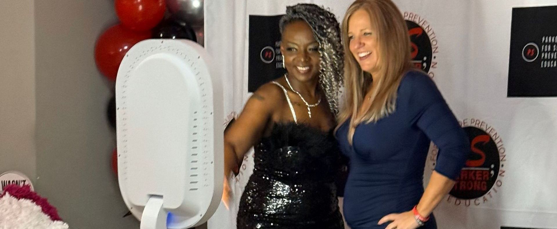 Two women smiling, posing at a photo booth with red and black balloons in the background.