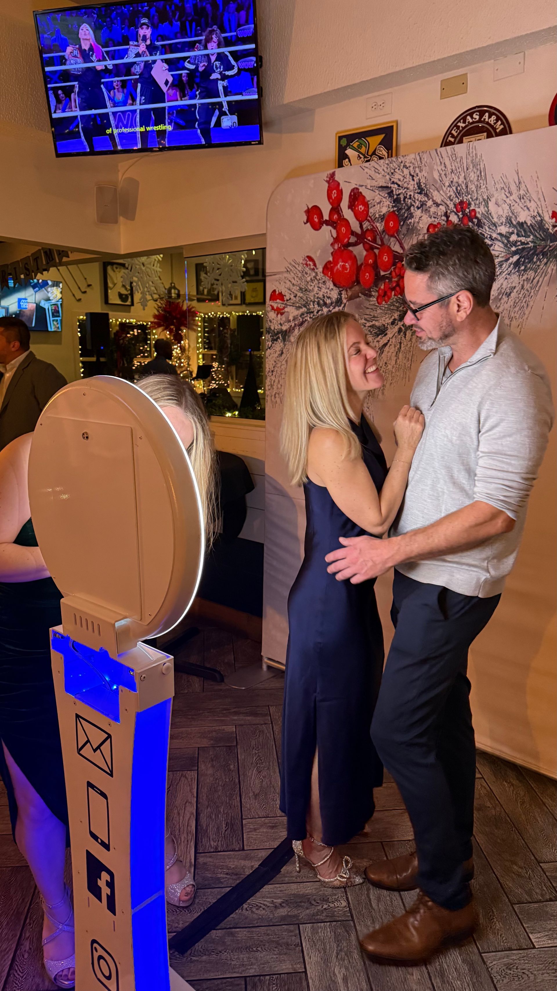 Couple poses for a photo near a photo booth, both smiling, inside a venue with a TV and decorations.