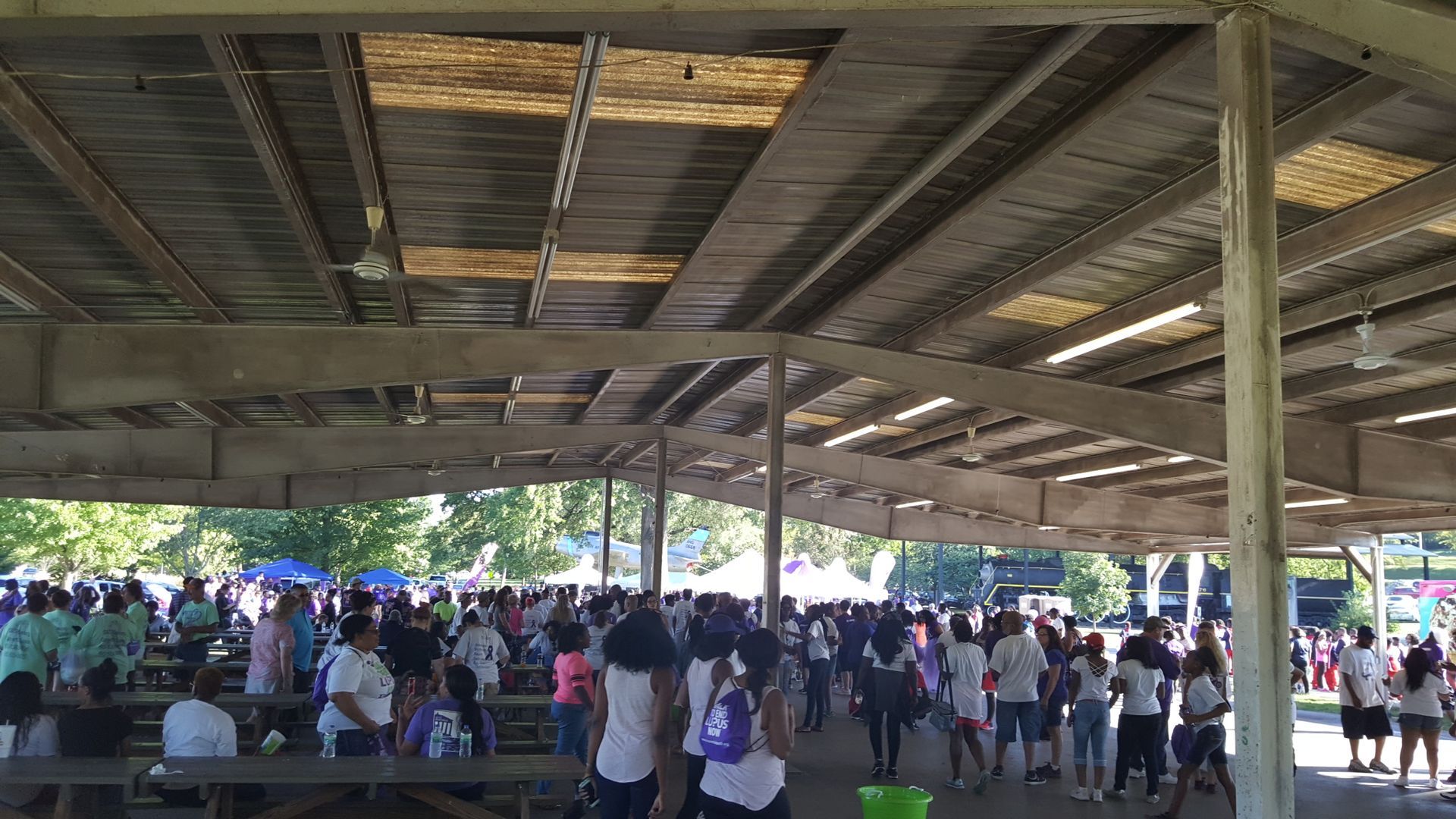 Large group of people gathered under a pavilion. Many wear white shirts. Sunlight streams through the roof.
