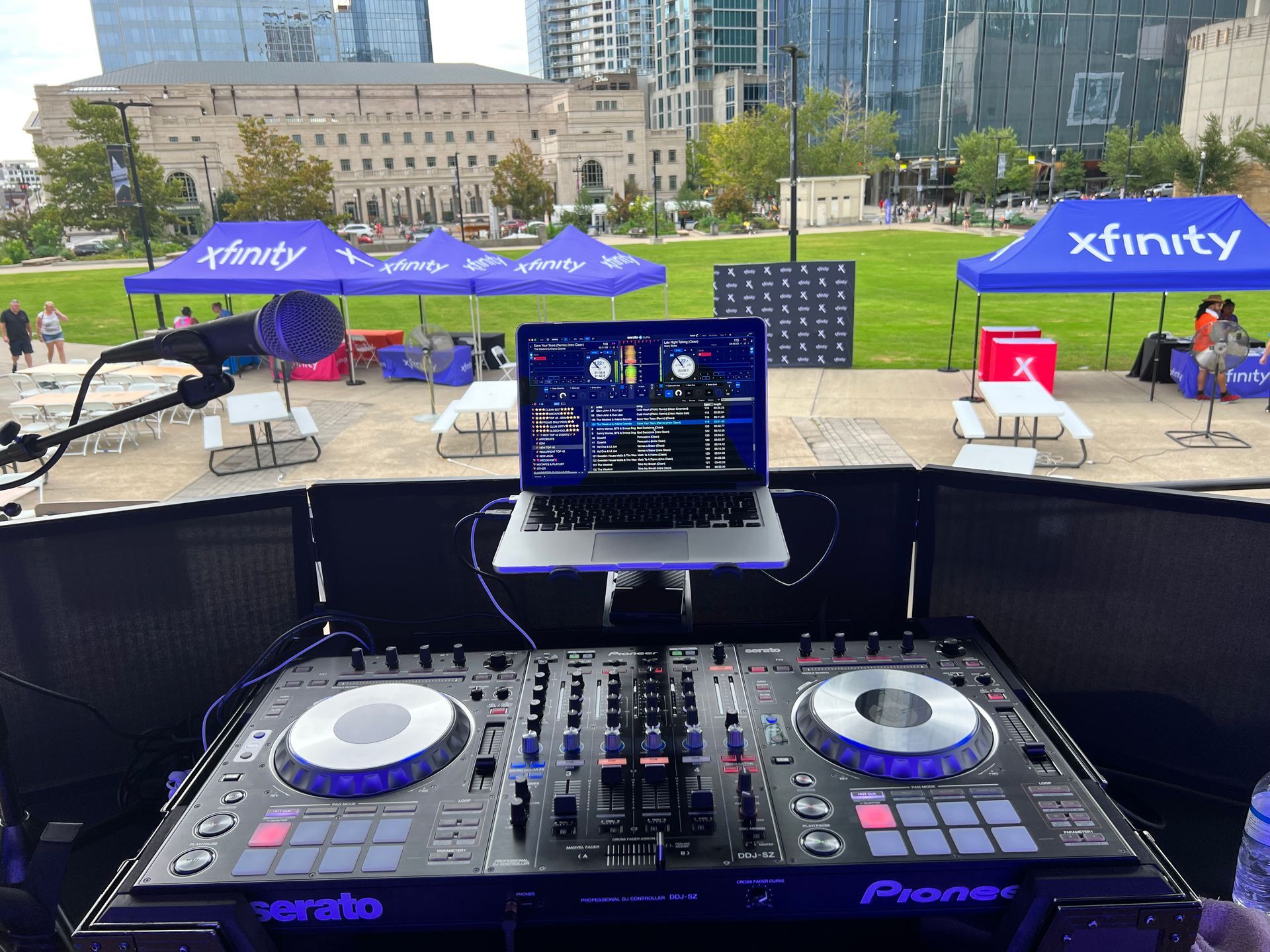 DJ setup outdoors with laptop, mixer, and microphone; Xfinity tents in background.