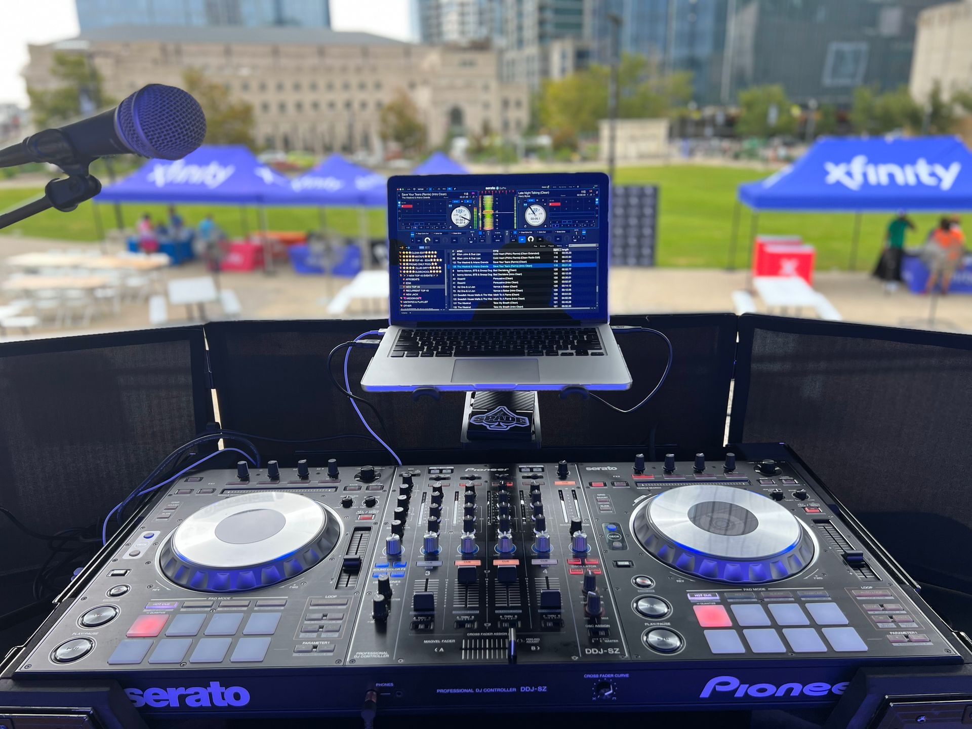 DJ setup outdoors with laptop, Pioneer controller, microphone, and Xfinity tent in the background.
