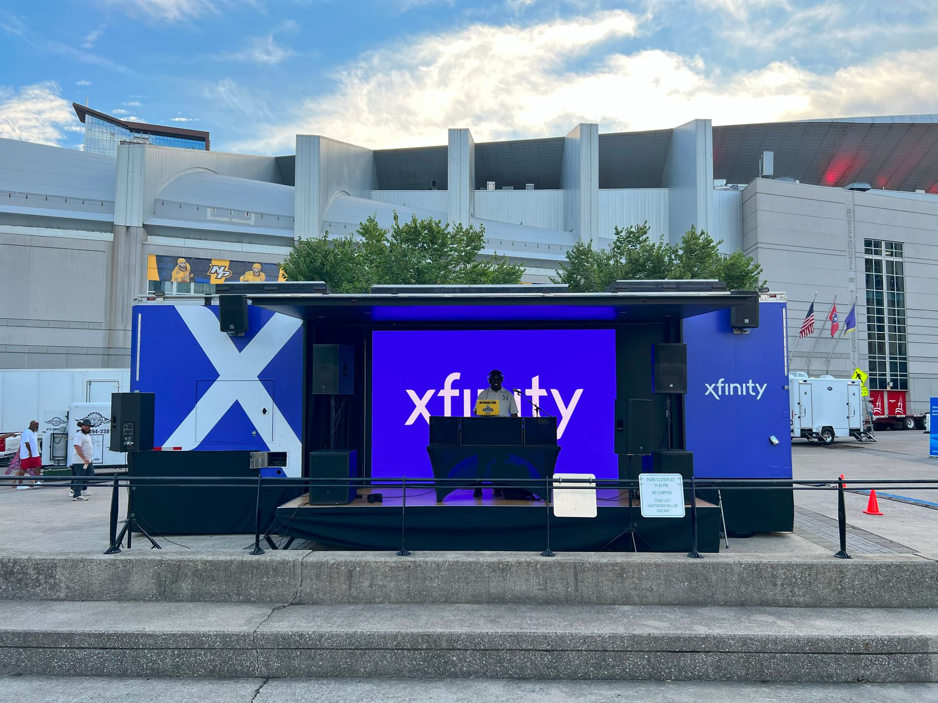 Xfinity stage outdoors; DJ at console, blue backdrop with logo, speakers, in front of a stadium.