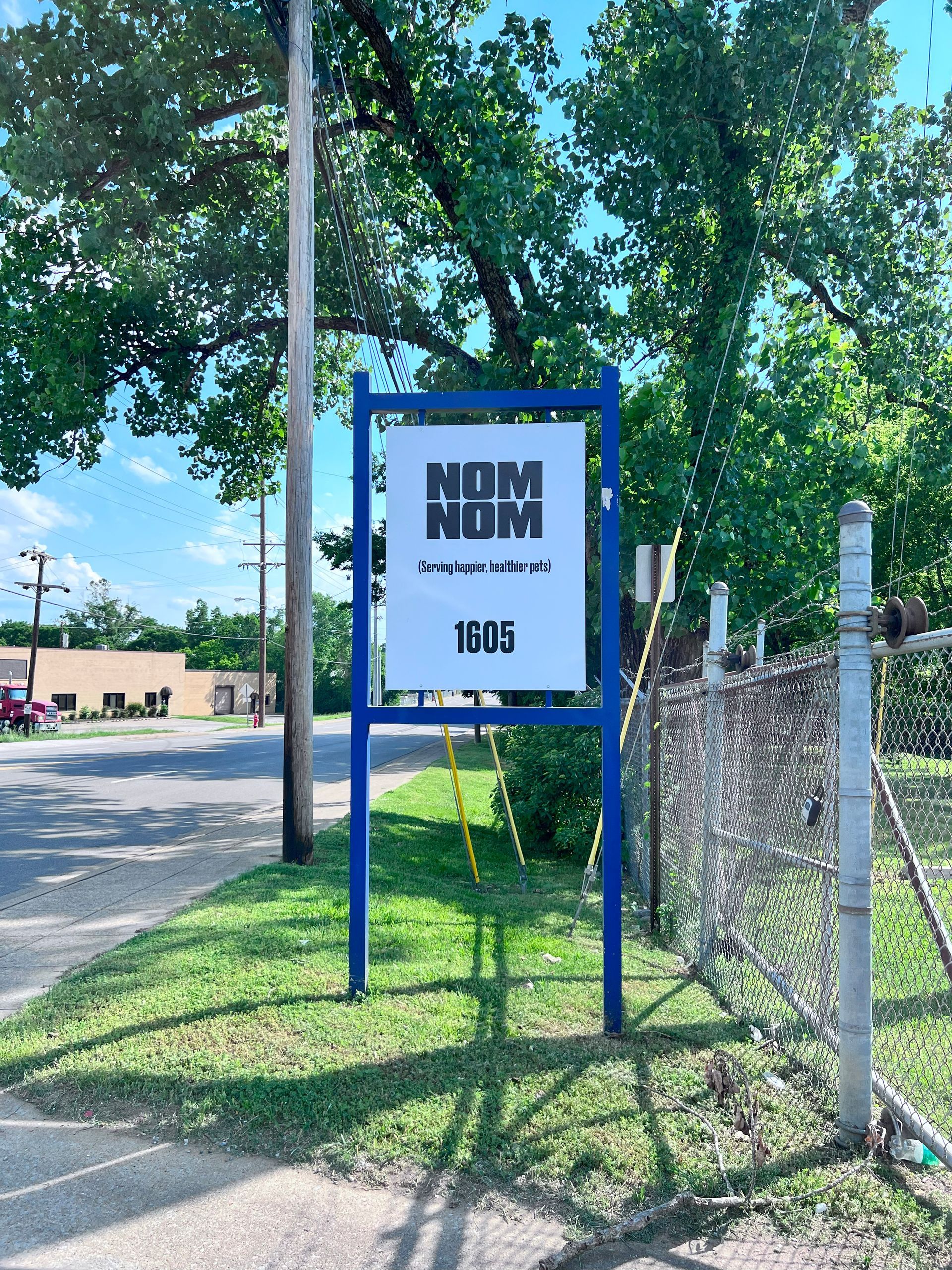 Blue sign: NOM NOM, with address 1935, on a grassy patch beside a chain-link fence.