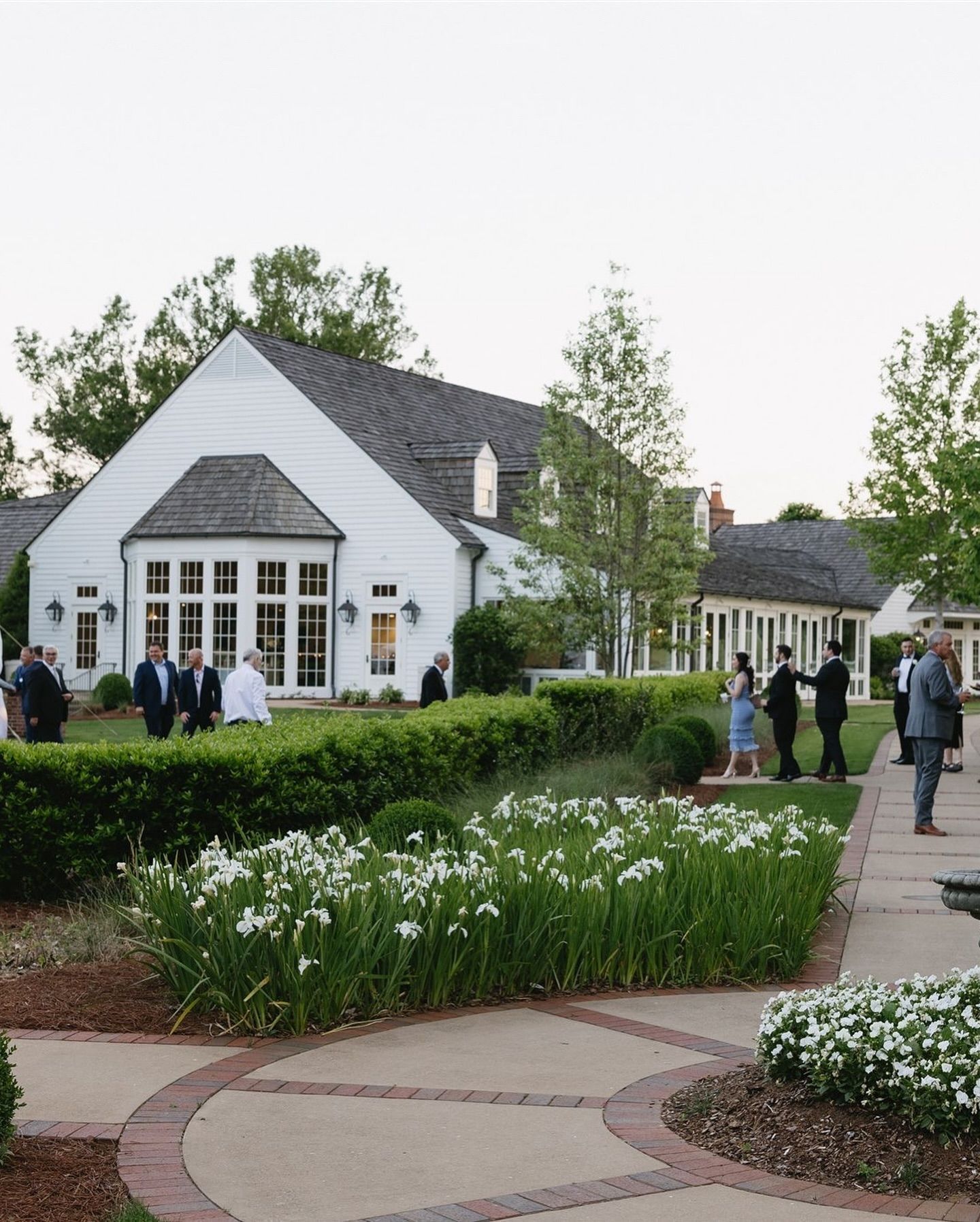 White building with a gray roof and several people gathered outside on a lawn. A flower garden is in the foreground.