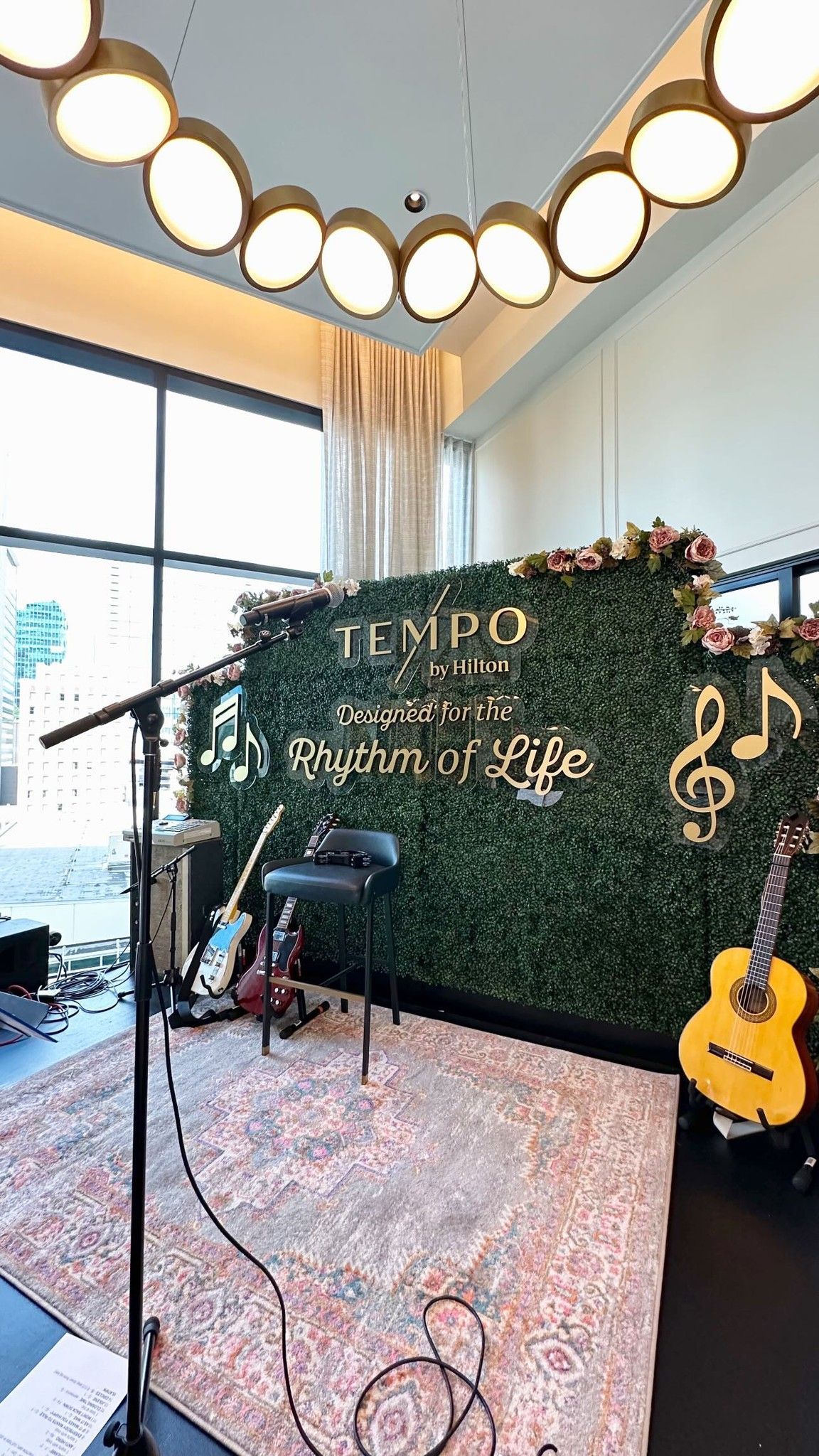 Live music setup: microphone, guitars, and stool in front of a greenery wall with event title and musical notes.