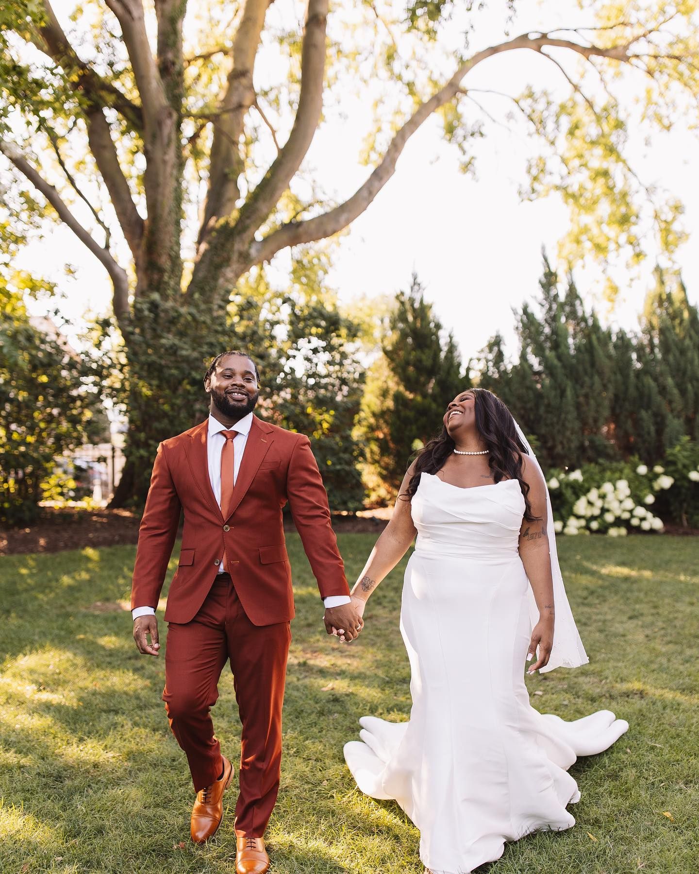 Couple in wedding attire hold hands and walk on grass, sunlight through trees.