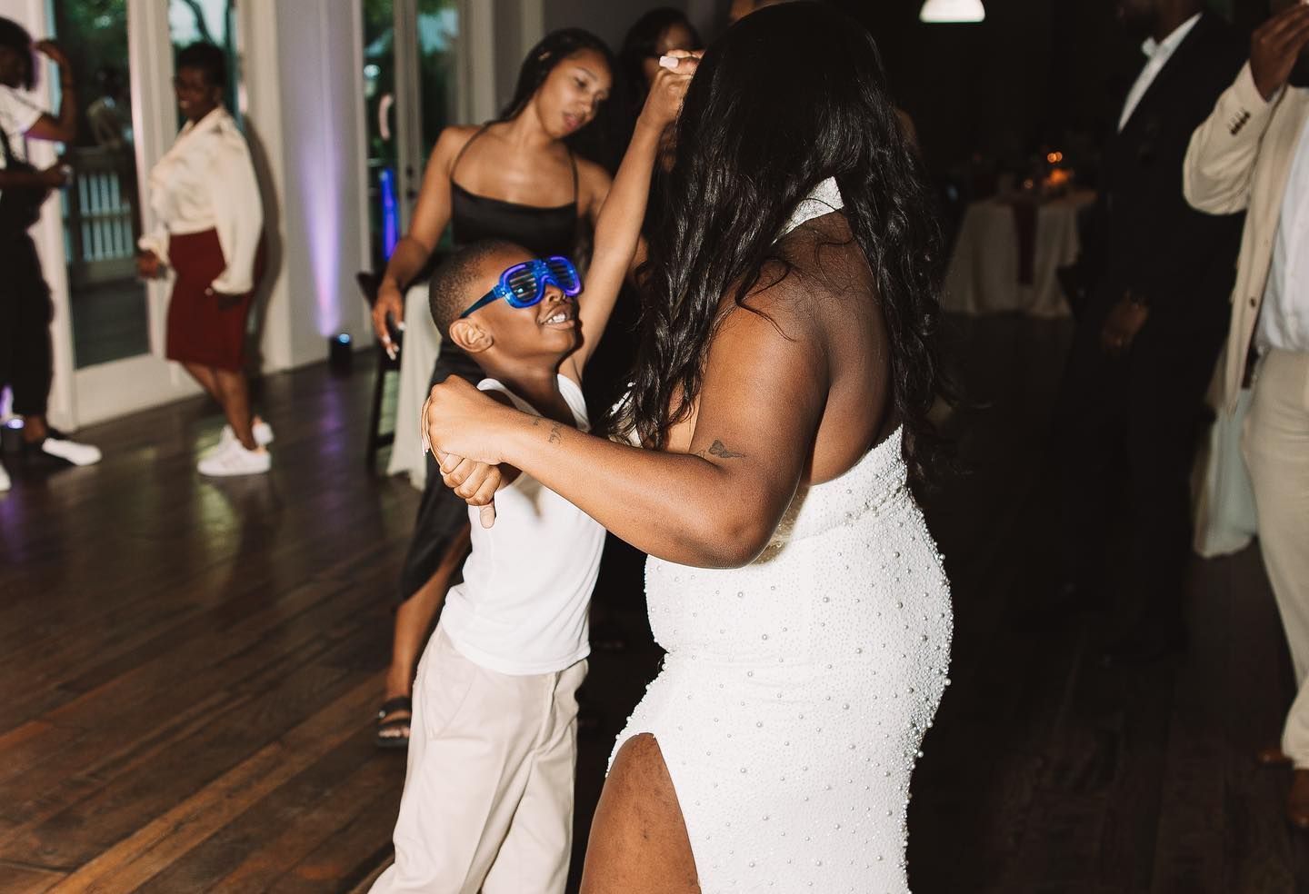 Woman in white dress dances with a child wearing blue toy glasses on a wooden dance floor.