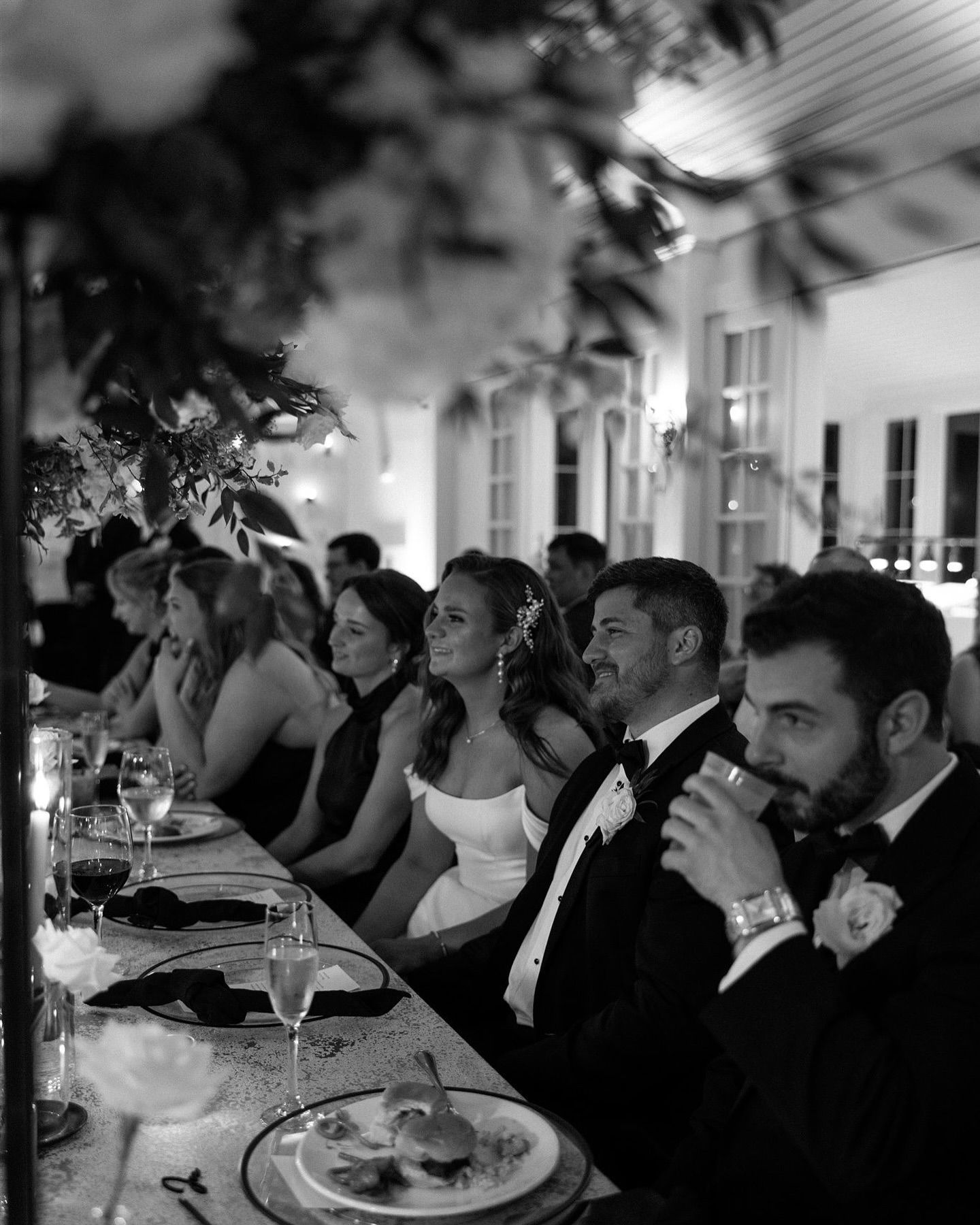 Wedding reception guests seated at a long table. Bride smiles, others look on. Black and white.