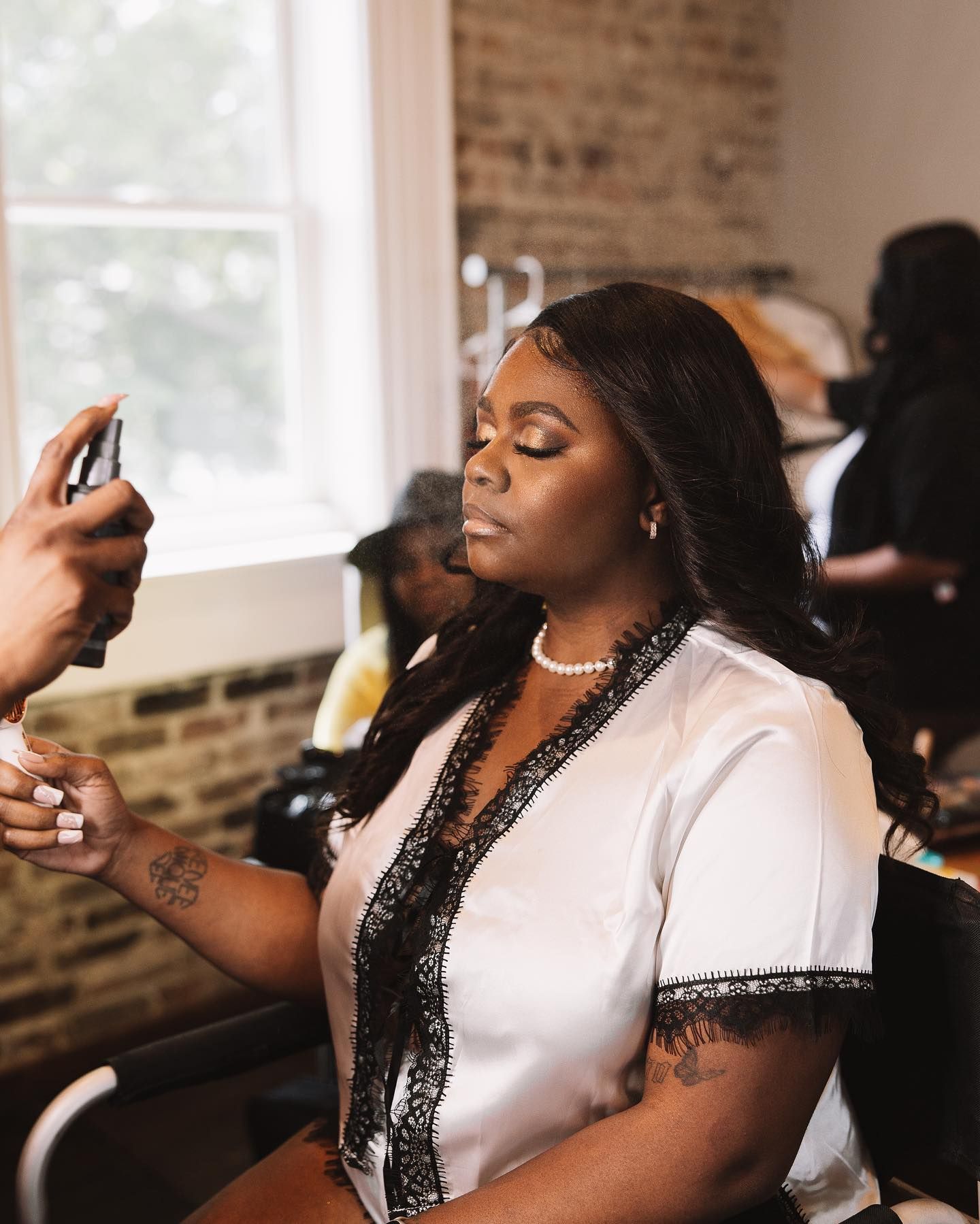 Person in white robe getting makeup sprayed in a bright room with a brick wall.