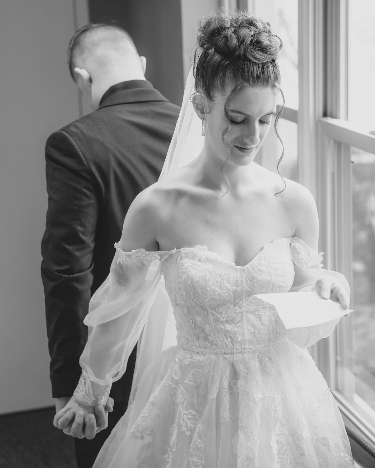 Bride in a wedding gown holding a letter, looking down. Groom stands behind, hand on hers.