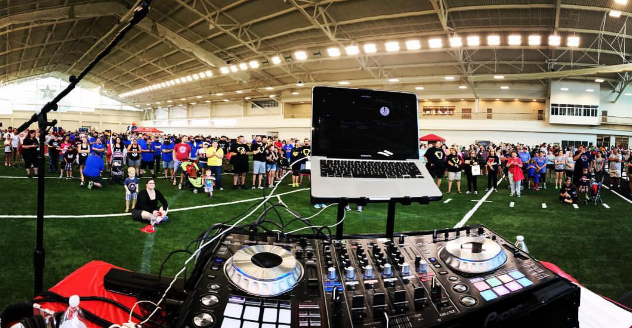 DJ setup with laptop and mixer, playing for a large crowd on an indoor sports field.