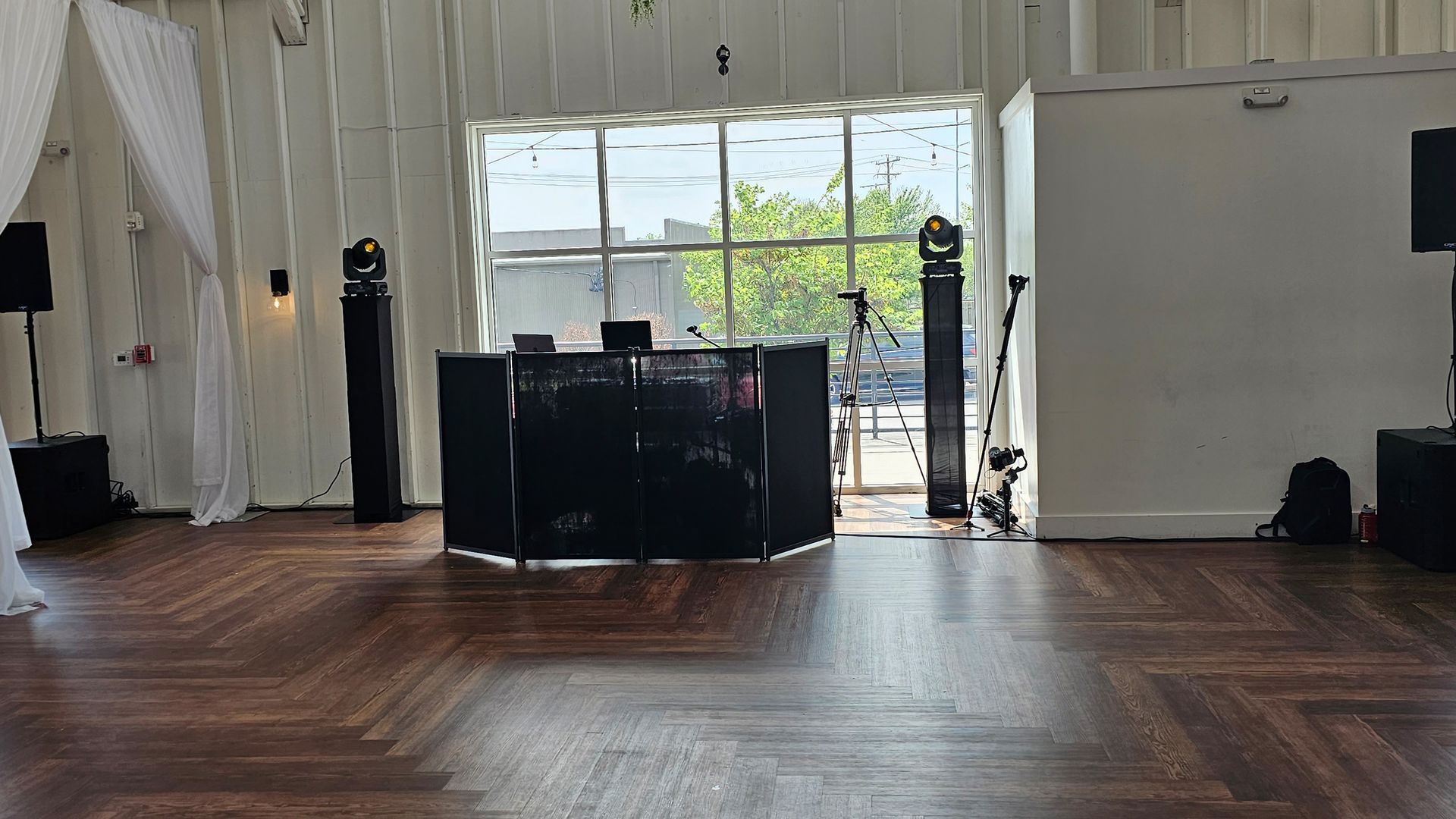 DJ setup in a room with hardwood floors and large window. Speakers and lights flank a black DJ booth.