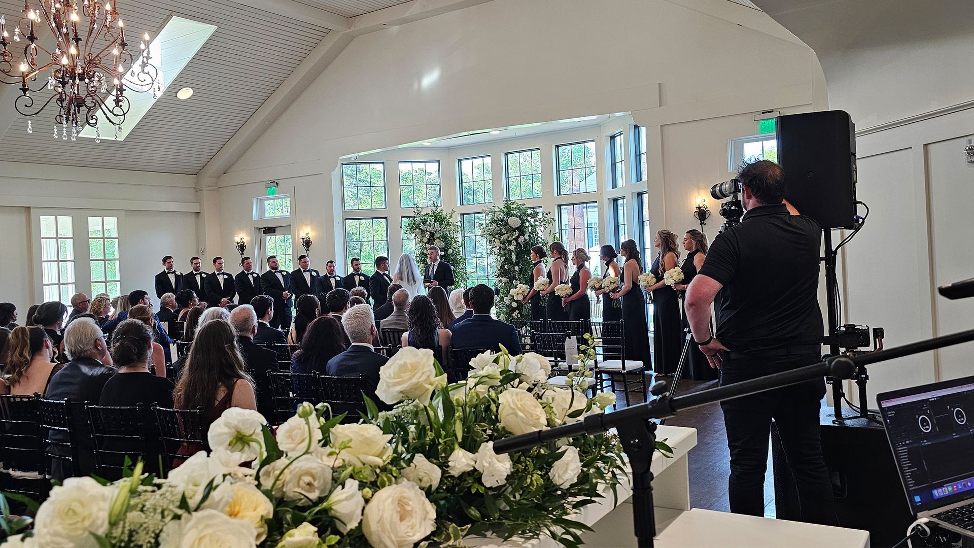 Wedding ceremony with guests, couple, and bridal party in a light-filled venue with floral decorations and audio equipment.