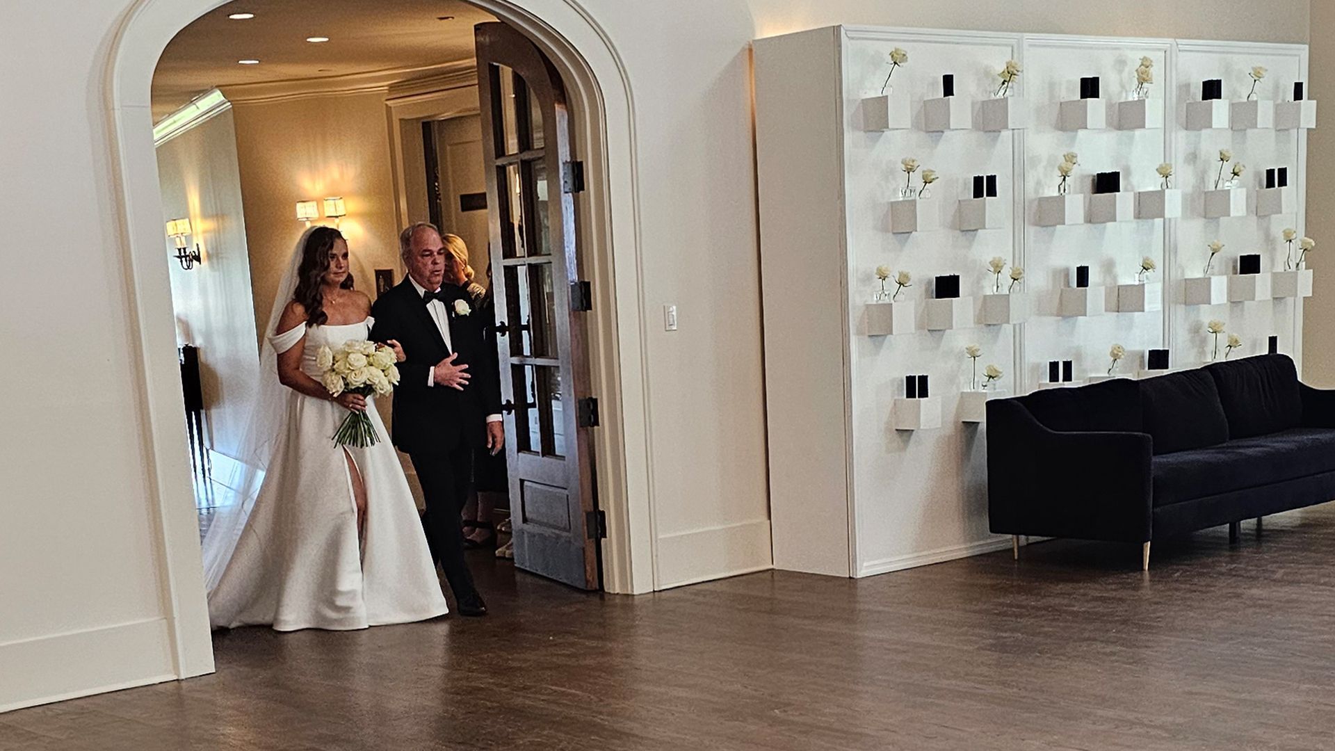 Bride in white gown walks with a man in a tuxedo through a doorway into a reception area with a wall display.