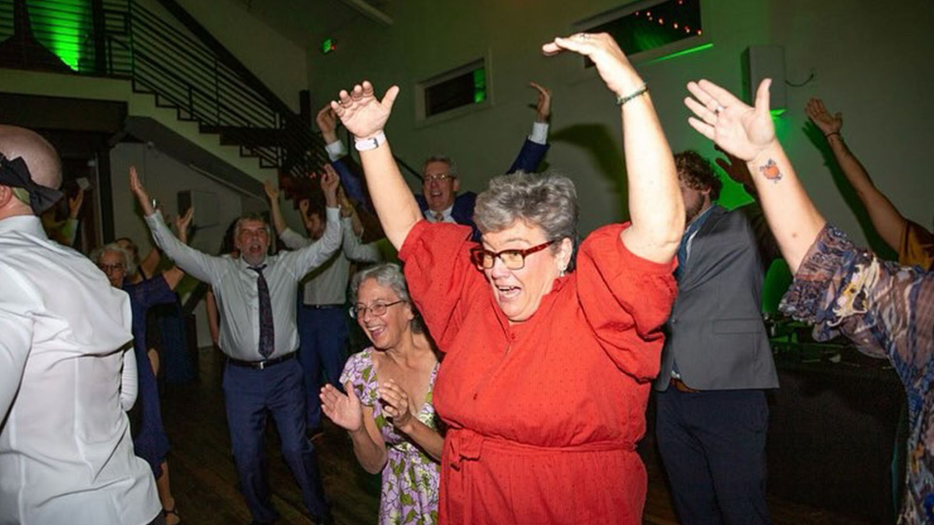 People dancing with arms raised at an event. Woman in red dress cheers with others. Green light shines.