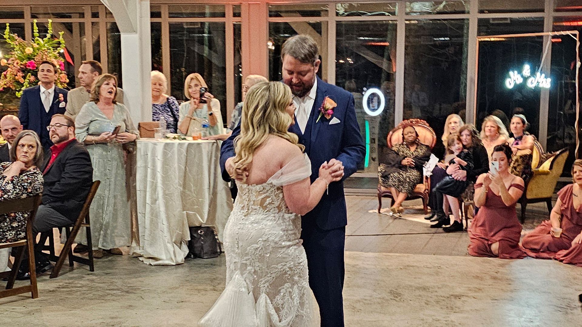 Couple dancing at wedding reception. Guests watch, some clapping. Indoors, lit venue with tables, flowers.