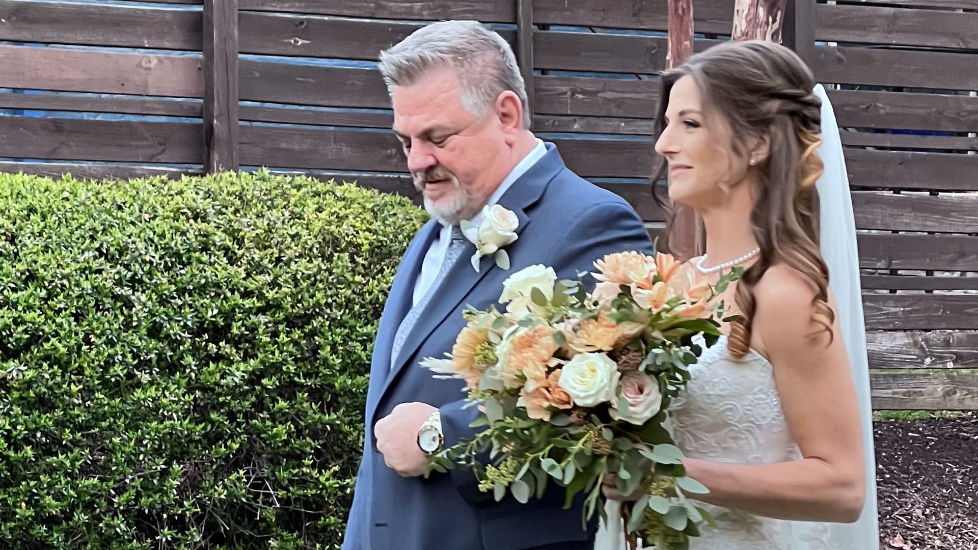 Bride and her father walk down an aisle, she carries a bouquet, he wears a suit.