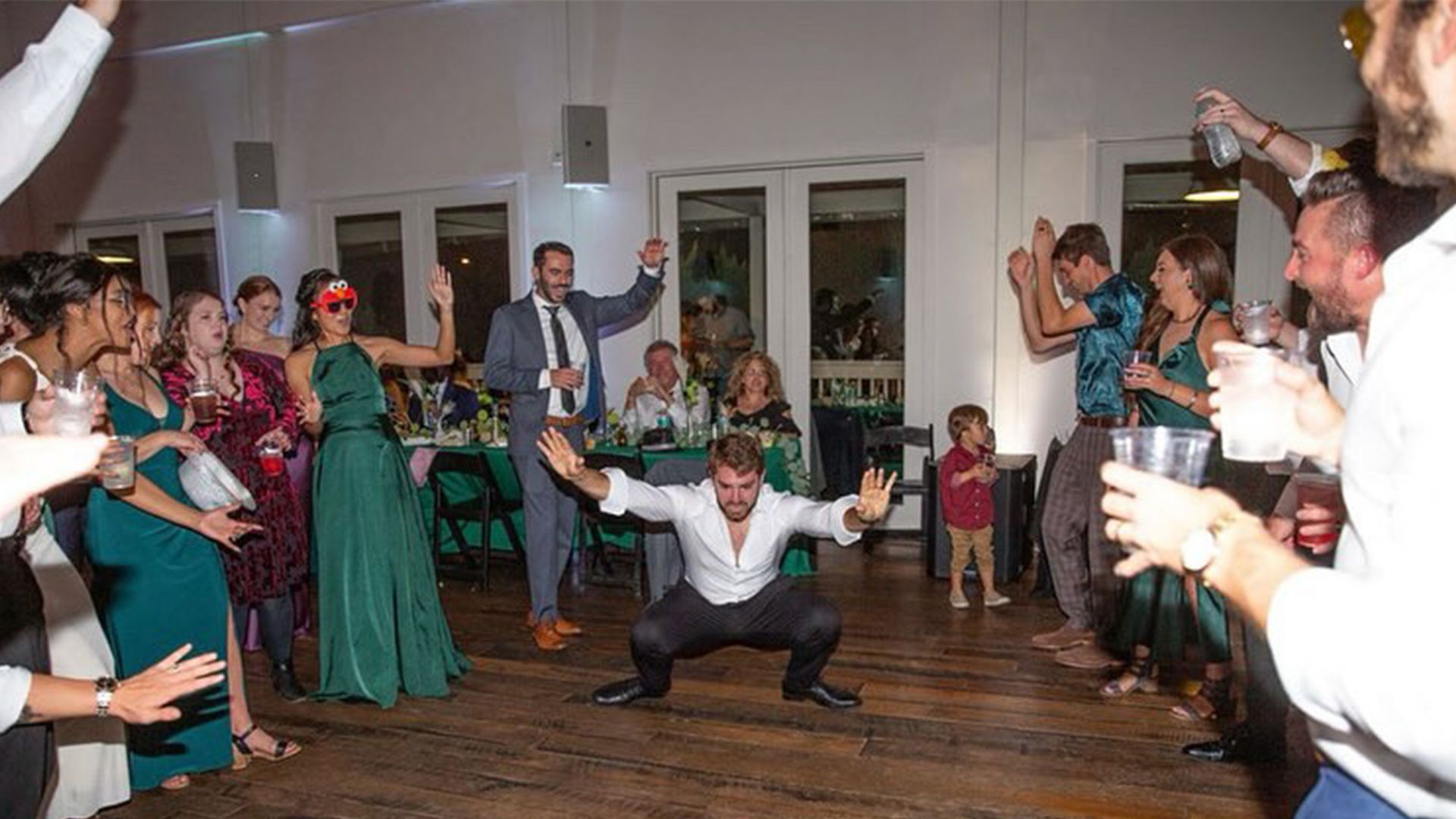 People dancing at a party; man in center doing a squat dance, others raise arms, festive setting.