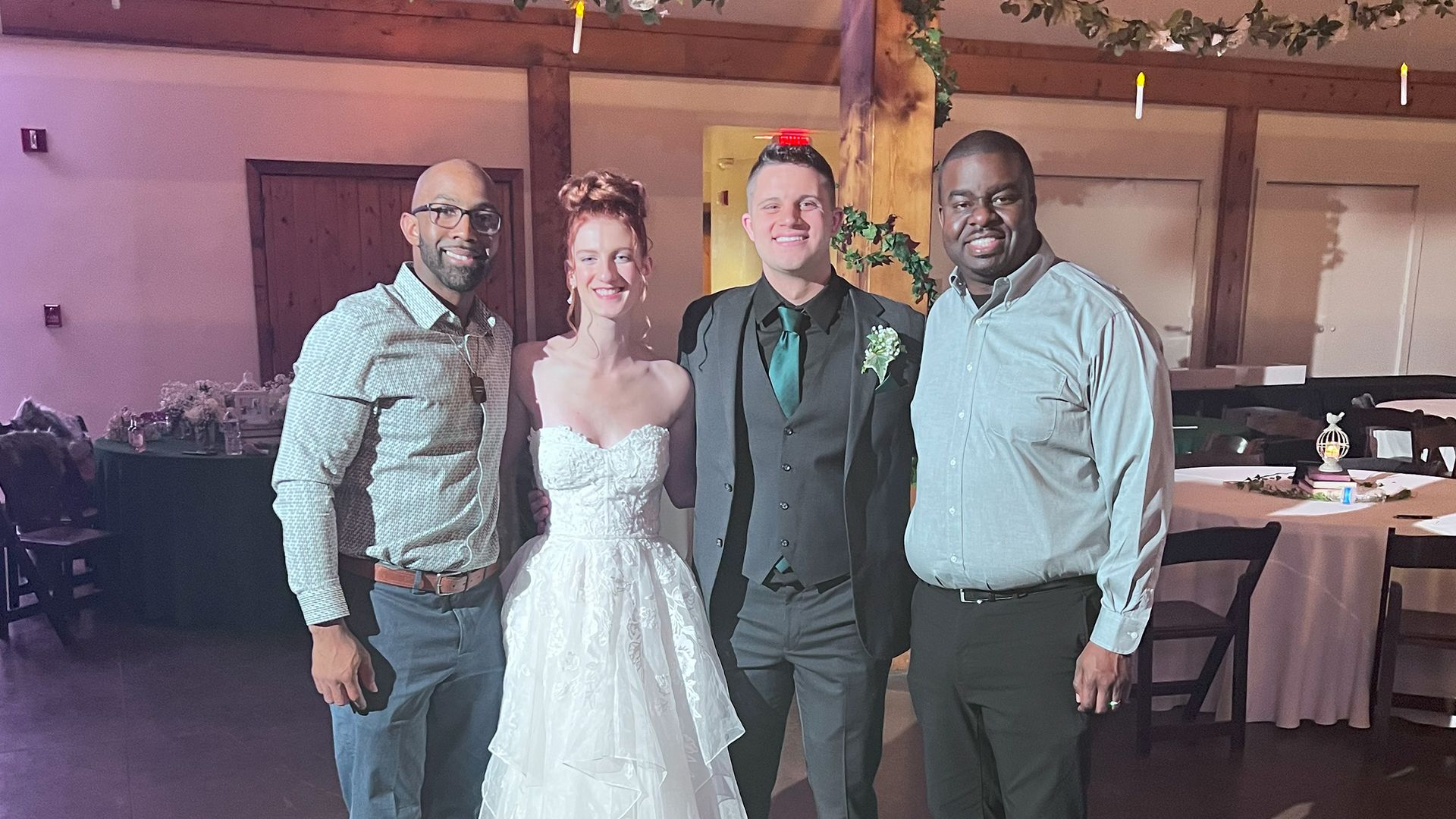 Four people pose indoors: a bride in a strapless dress, groom in suit with teal tie, and two men, all smiling.