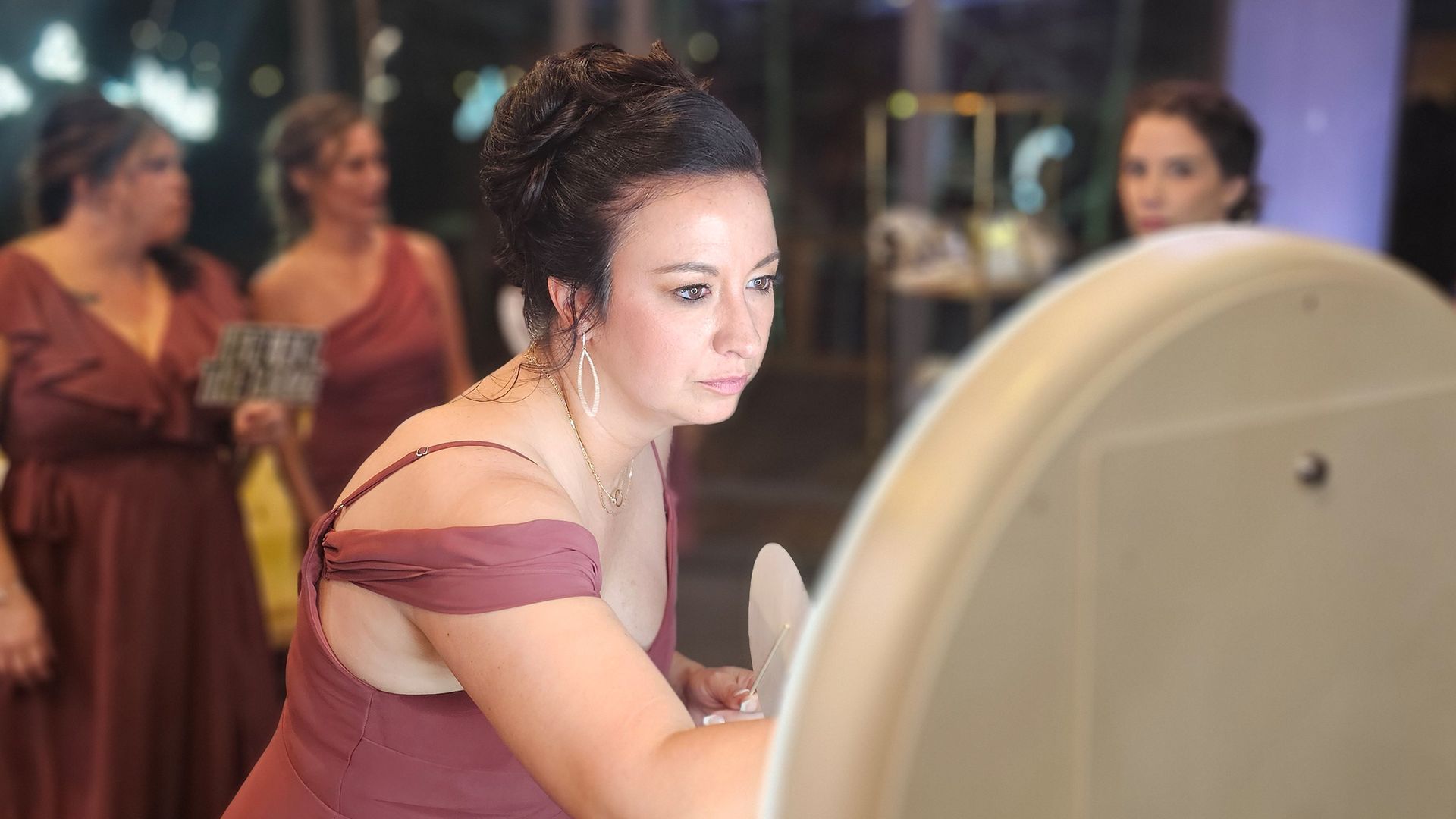 Woman in a mauve dress using a photo booth, three others in similar dresses stand behind her at an event.
