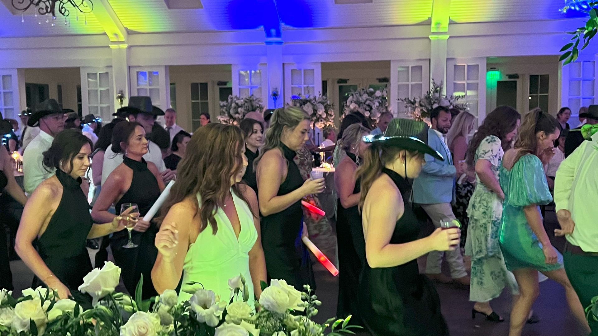 People dancing at an outdoor event with lights. Some wear cowboy hats, holding drinks, in front of flowers and buildings.