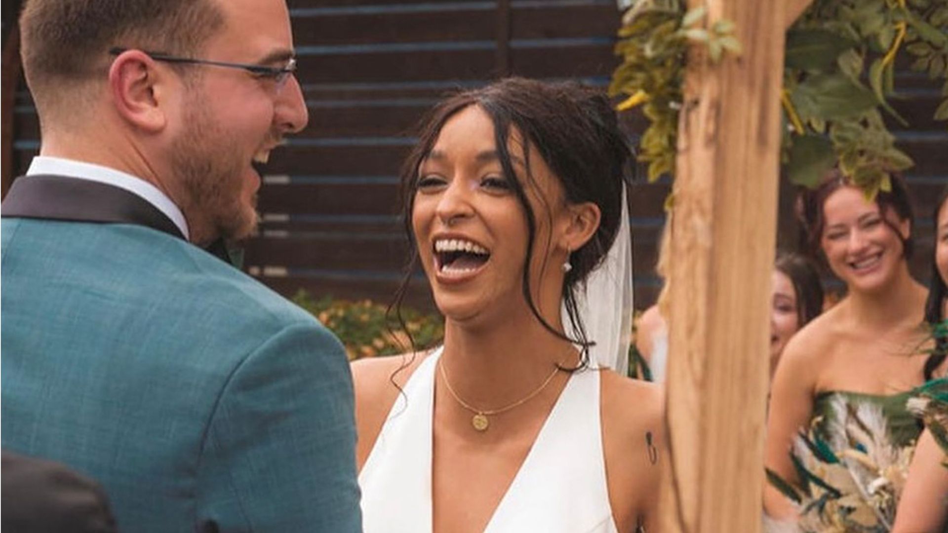 Bride and groom laugh during wedding ceremony. They stand under a wooden archway outdoors. Green plants and guests are visible.