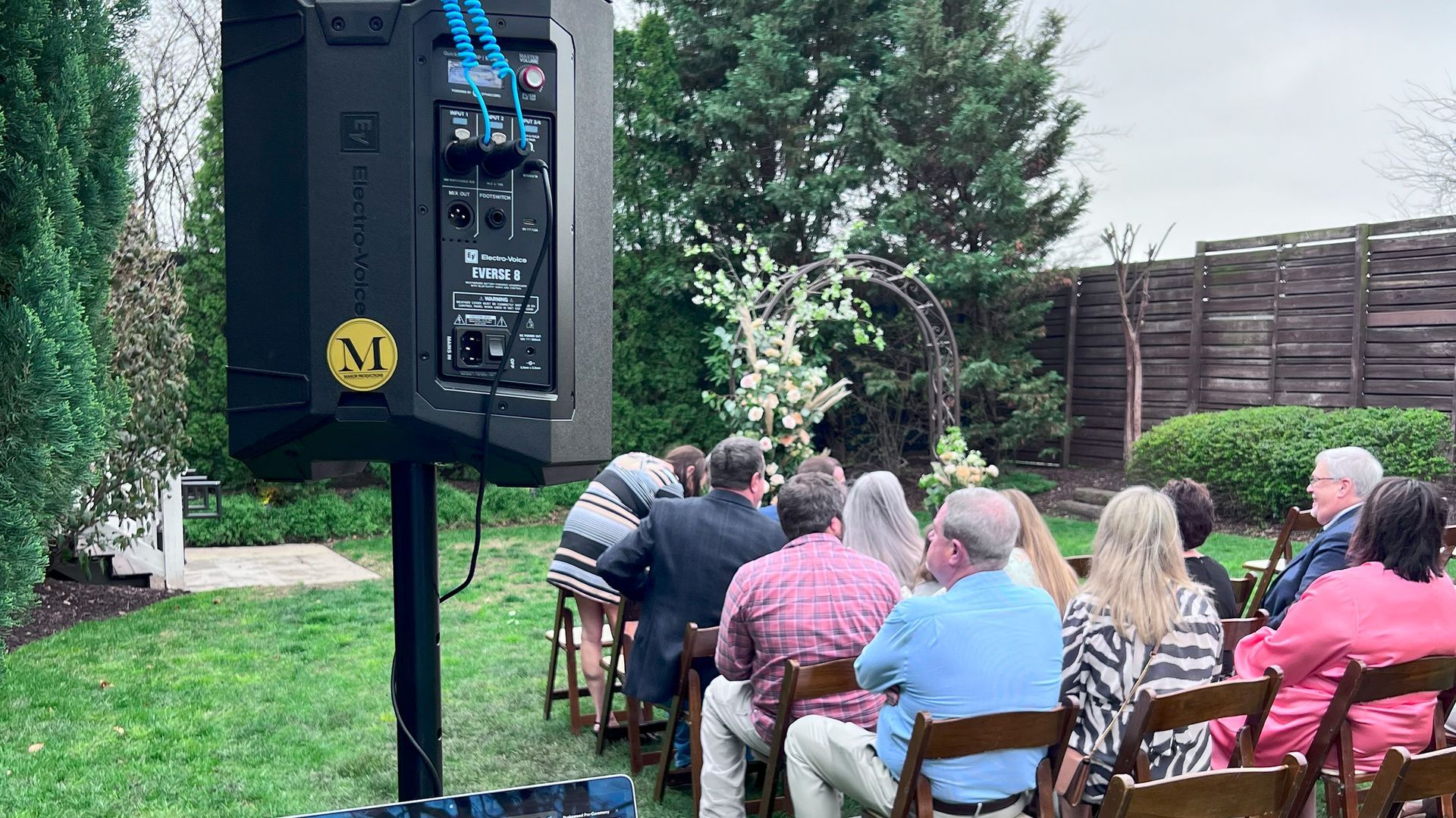 Outdoor wedding ceremony with guests seated, a speaker, and an arch.
