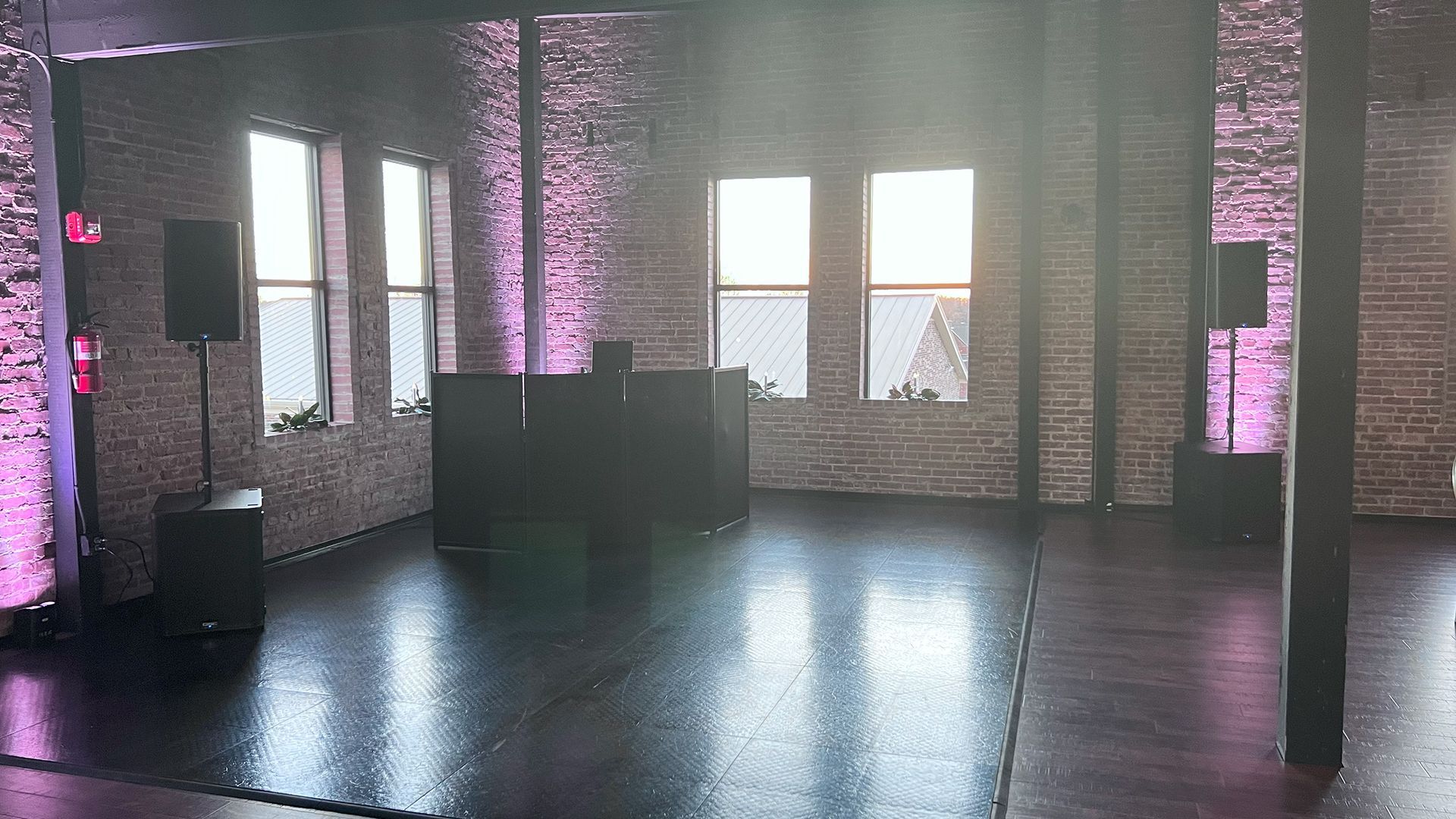 Empty event space with brick walls, windows, DJ setup, and stage lighting in purple.