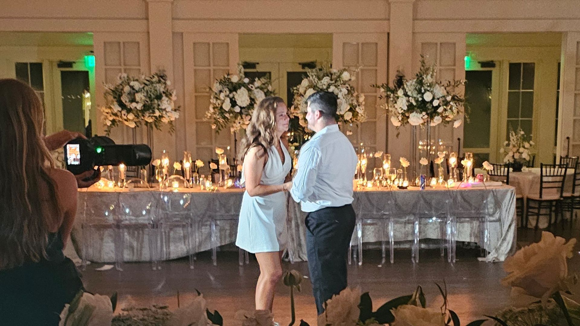 Couple facing each other with a photographer present, lit table with flowers and candles in a ballroom setting.