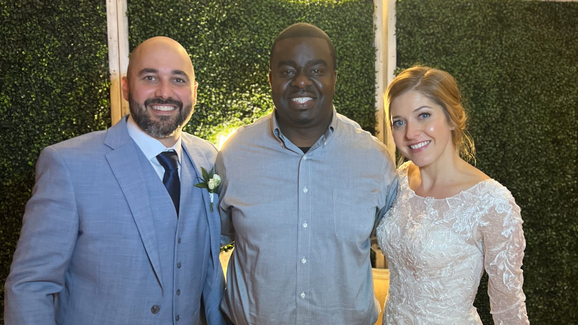 Three people smiling for the camera in front of a green hedge; a man in a blue suit, a man in a gray shirt, and a woman in a wedding dress.