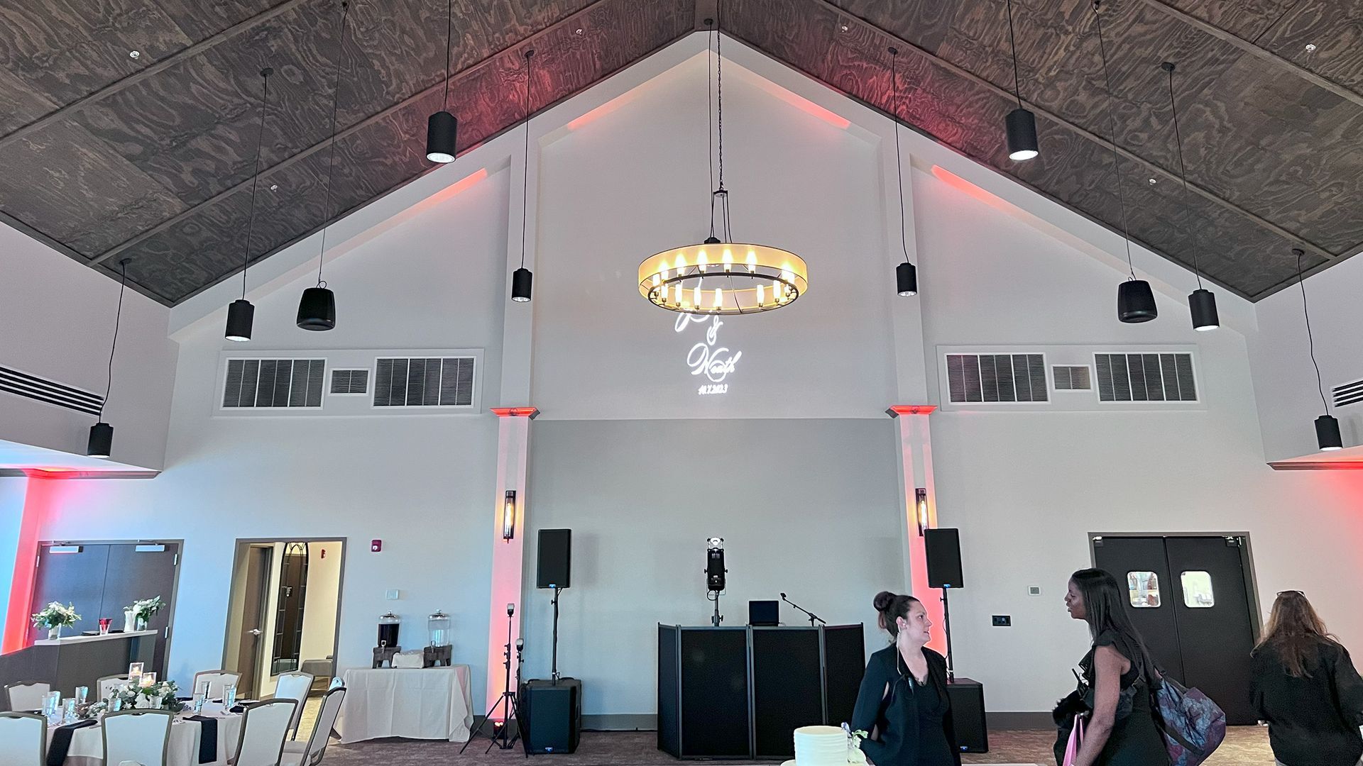 Wedding reception hall, high ceilings, red lighting, DJ equipment set up, guests milling about.
