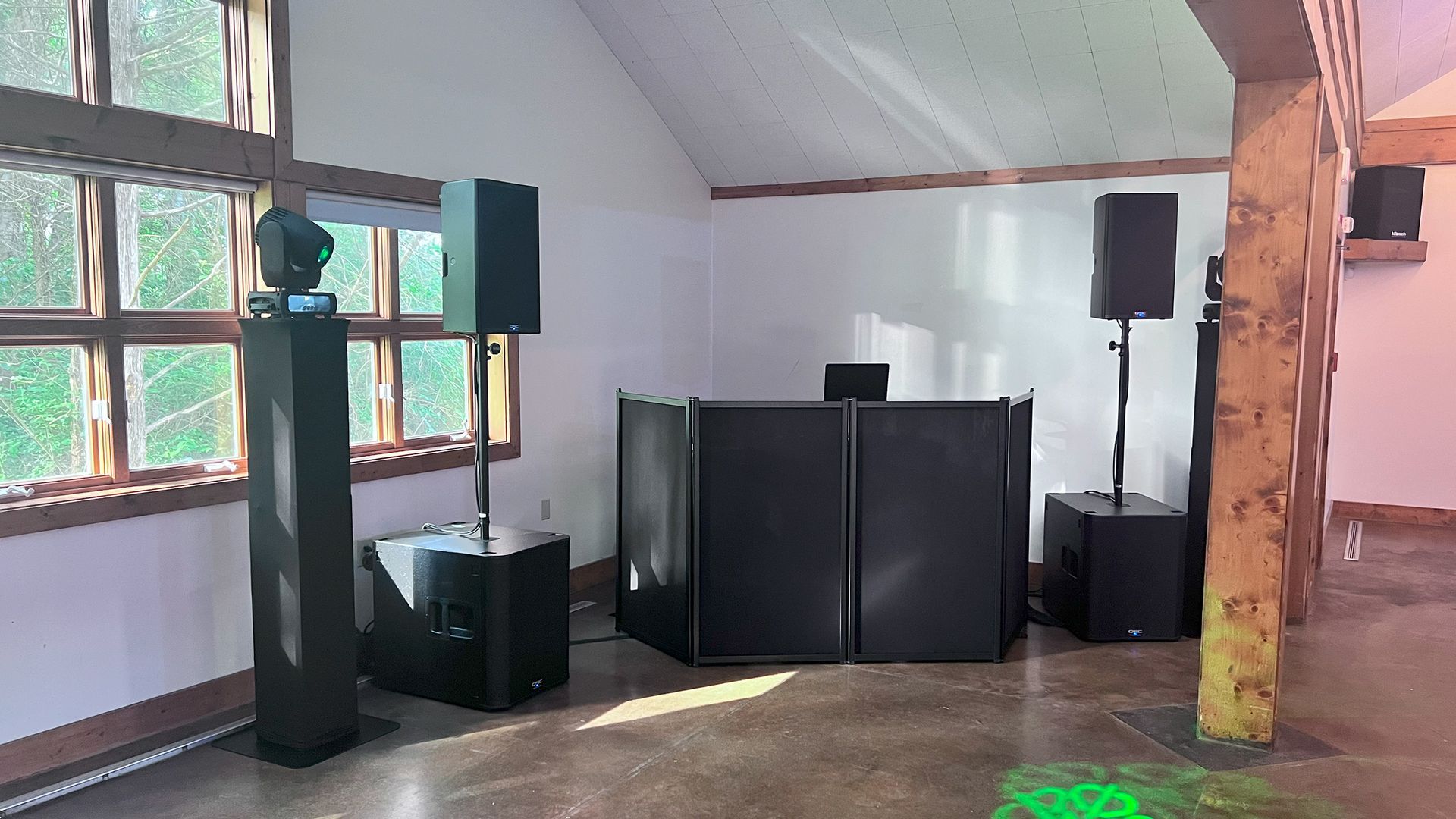 DJ setup with speakers, lights, and a booth in a room with a large window and wooden beams.