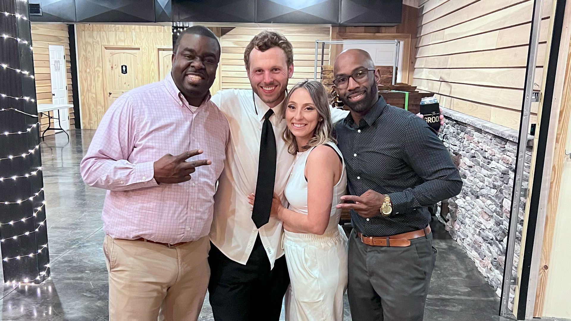 Four people posing together, likely at a celebration. Smiling and giving peace signs. Indoors, rustic setting.