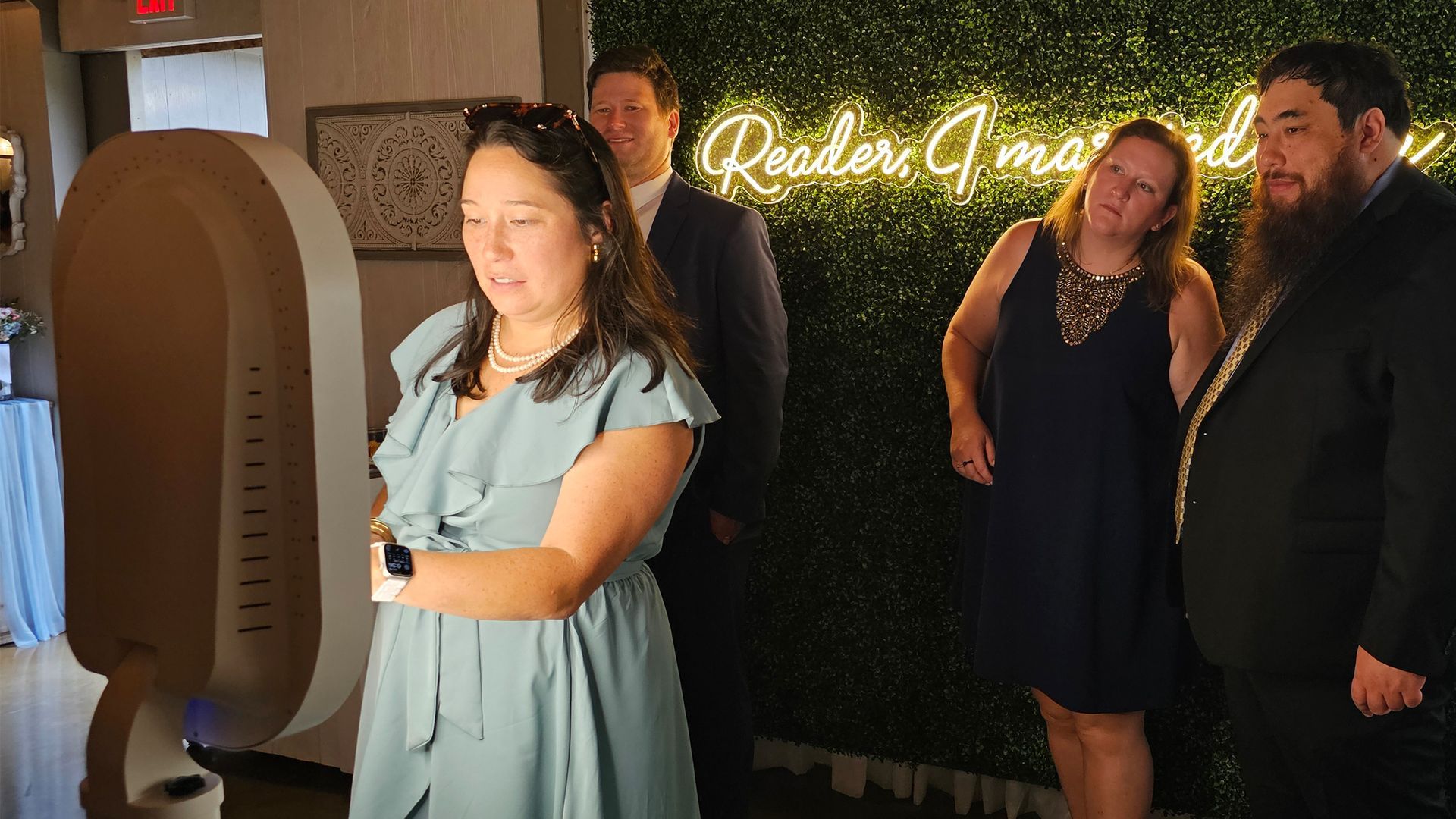 People posing at a photo booth with green wall backdrop, neon sign. One woman looking at the camera with others behind.